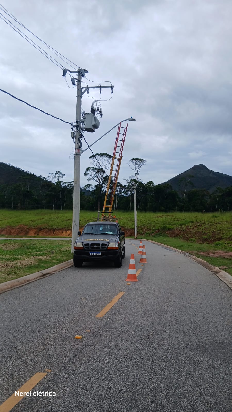 Escada apoiada em poste de iluminação em via com pista asfaltada, montanhas ao fundo e caminhonete da Nerel Elétrica sinalizada com cones