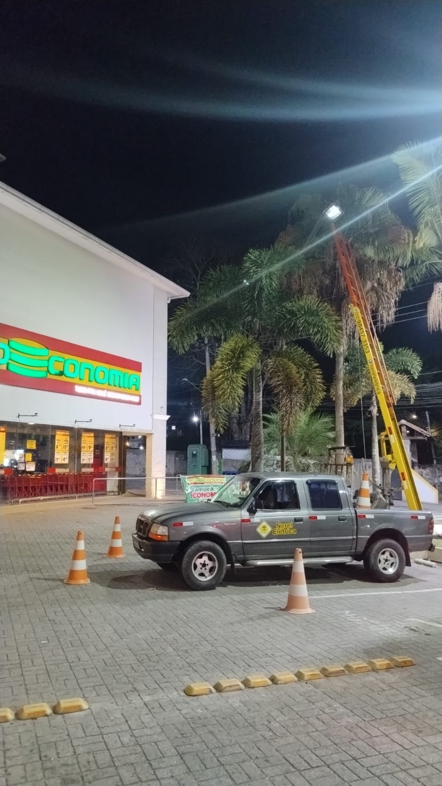 Atendimento noturno em área comercial com refletor instalado no alto e equipe da Nerel Elétrica com caminhonete e cones no estacionamento
