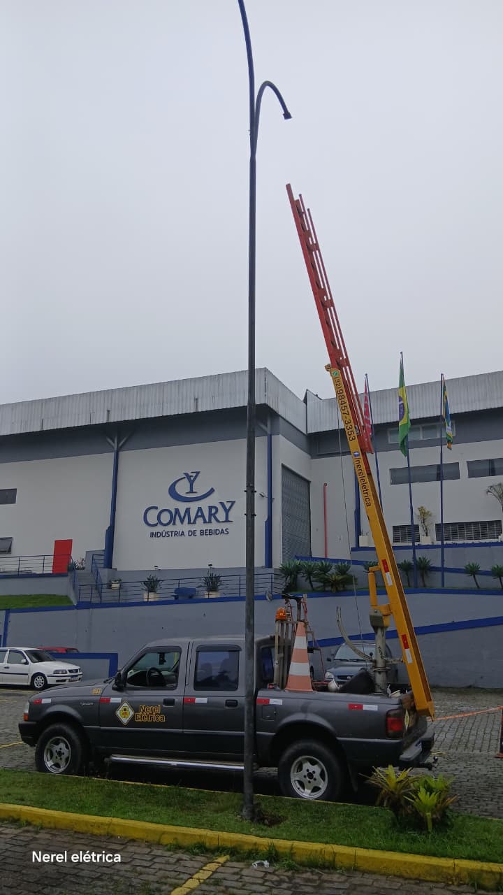 Poste de iluminação em frente a galpão industrial com escada da Nerel Elétrica apoiada e caminhonete estacionada